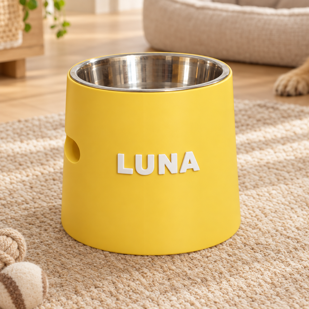 A yellow pet feeder bowl with a stainless steel interior and the name 'LUNA' in white letters on the front, placed on a beige carpet in a home setting.