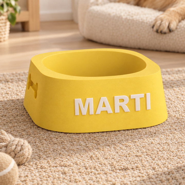 A bright yellow pet feeder bowl with the name 'MARTI' in white raised letters on the front, placed on a beige carpet in a home setting with a dog's paw visible in the background.